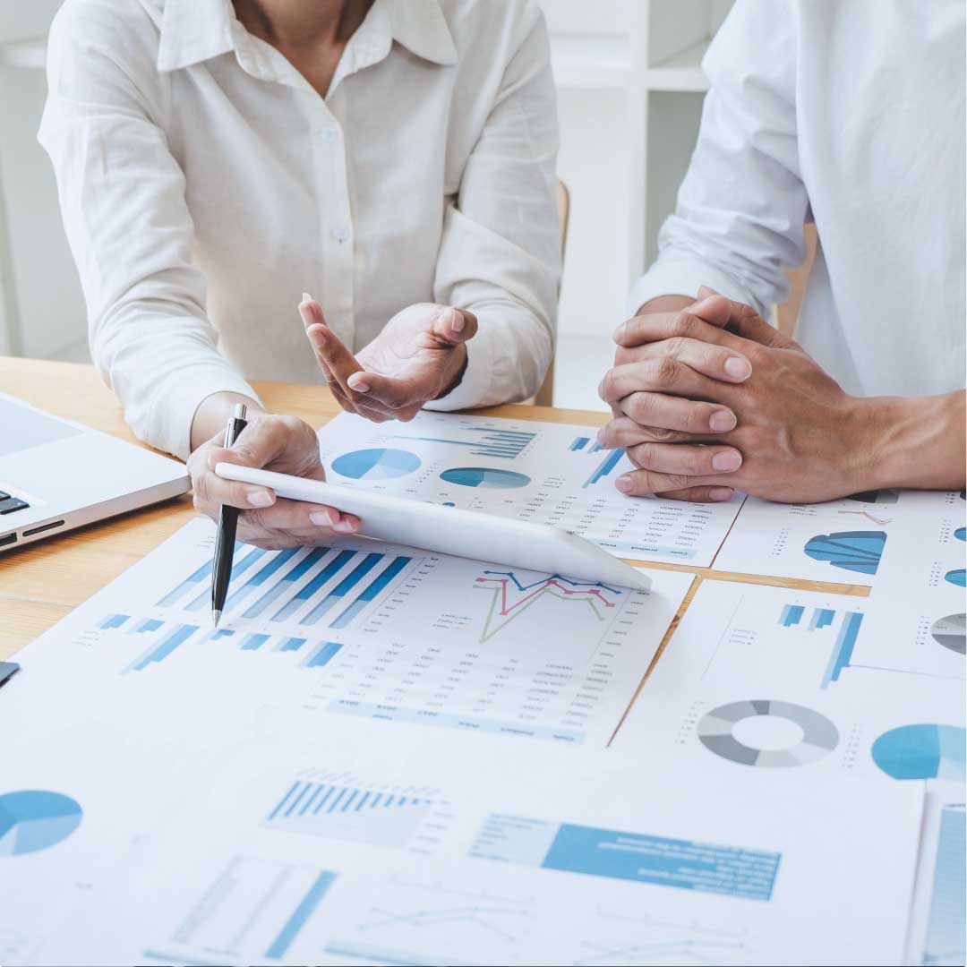 A woman is seated next to a man, explaining a financial situation using hand gestures and printed documents that display various types of graphs.